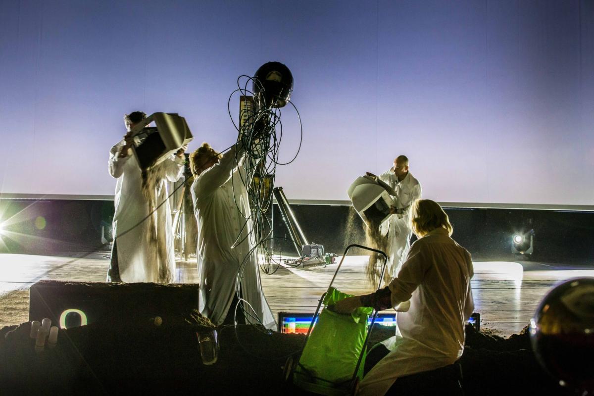 Das FARN Kollektiv während der Aufführung von "The Shape of Trouble to Come" in weißen Kitteln, mit alten technischen Geräten und Erde auf einer Bühne