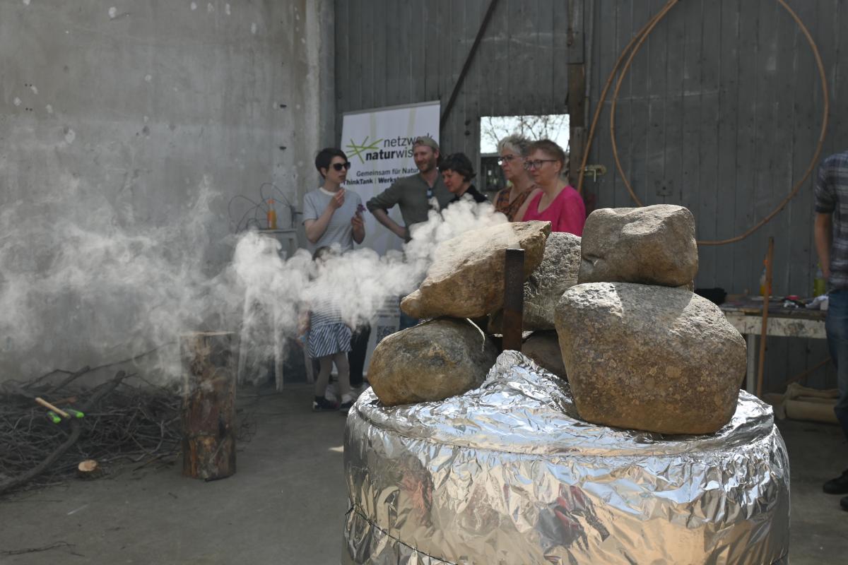 Eine Köhlertonne steht im Atelier der Künstlerin, der Deckel ist mit Steinen beschwert, aus einem Rohr steigt heller Rauch auf.