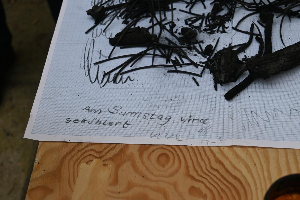 Auf Millimeterpapier liegen einige verkohlte Pflanzenfragmente. Auf dem Papier steht in Kohle geschrieben "Am Samstag wird geköhlert".