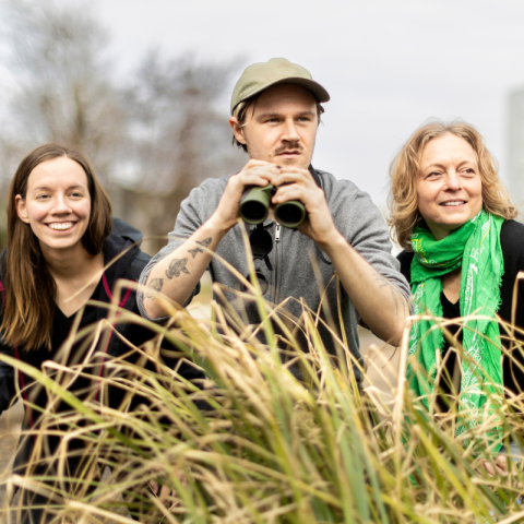 Drei Personen im Vordergrund mit einem Ferngläsern und mehreren Pflanzen um das Trio. 